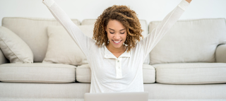 Eine fröhliche Frau mit lockigem Haar sitzt auf dem Boden vor einem Laptop und hebt die Arme in Freude. Im Hintergrund sieht man ein helles Sofa.