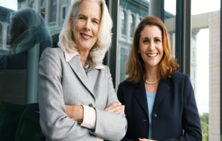 Das Bild zeigt zwei lächelnde Frauen in eleganter Geschäftskleidung, die vor großen Fenstern stehen. Sie strahlen Selbstbewusstsein aus.