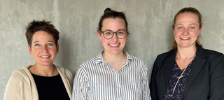 Das Bild zeigt drei Frauen, die lächelnd vor einer neutralen, grauen Wand stehen. Sie tragen legere und professionelle Kleidung.