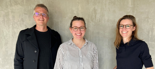 Das Bild zeigt drei Personen, die freundlich lächeln. Sie stehen vor einer grauen wand und tragen lässige Kleidung, die Brillen betonen ihren Look.