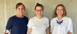 Das Bild zeigt drei Frauen, die freundlich in die Kamera lächeln. Sie stehen vor einer neutralen Wand und tragen verschiedene, moderne Outfits.