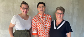 Drei Frauen stehen nebeneinander vor einem grauen, schlichten Hintergrund. Sie lächeln und tragen unterschiedliche, stilvolle Kleidung.