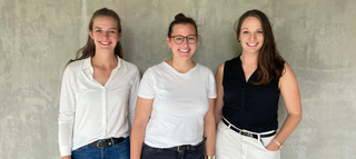 Drei Frauen stehen vor einer grauen Wand. Sie tragen verschiedene Kleidung und lächeln freundlich in die Kamera.