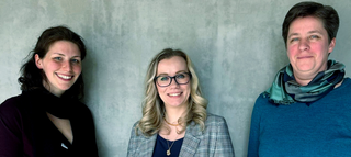 Drei Frauen stehen vor einer grauen Wand. Sie lächeln, tragen unterschiedliche Outfits und wirken freundlich und professionell.