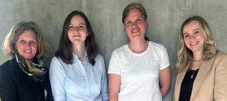 Vier Frauen stehen lächelnd nebeneinander vor einer grauen Wand. Sie strahlen Freundlichkeit und Teamgeist aus.