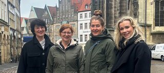 Vier Frauen stehen freundlich an einer historischen Stadtstraße. Im Hintergrund sind alte Gebäude und eine Kirche zu sehen. Ein bewölkter Tag.