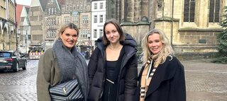 Drei Frauen stehen lächelnd in einer Stadtlandschaft mit historischen Gebäuden im Hintergrund. Es ist ein klarer Tag.