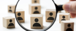 Das Bild zeigt Holzklötze mit menschlichen Symbolen. Eine Lupe vergrößert einen Block, was auf Fokus und Analyse hinweist.