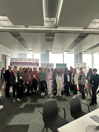 Das Bild zeigt eine Gruppe von Frauen und Männern, die an einer Veranstaltung zum Thema Mentoring teilnehmen. Sie stehen zusammen lächelnd in einem modernen Raum.
