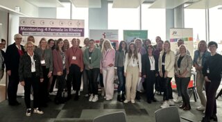 Das Bild zeigt eine Gruppe von Frauen und Männern, die an einer Veranstaltung zum Thema Mentoring teilnehmen. Sie stehen zusammen lächelnd in einem modernen Raum.