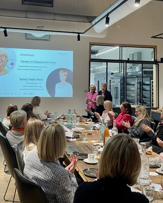 Das Bild zeigt eine Gruppe von Frauen, die applaudieren. Im Hintergrund präsentieren zwei Frauen bei einer Abschlussveranstaltung in einem modernen Raum.