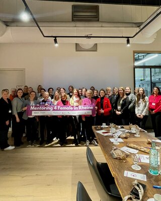 Das Bild zeigt eine Gruppe von Frauen, die zusammenstehen und ein Banner mit der Aufschrift "Mentoring 4 Female in Rheine" halten. Sie wirken freundlich und motiviert.