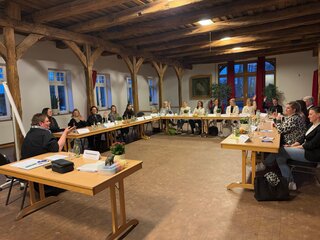 Das Bild zeigt eine Sitzung in einem gut beleuchteten Raum mit Holzbalkendecke. Teilnehmer*innen sitzen an Tischen, einige diskutieren aktiv.