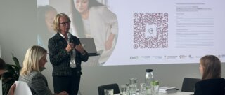 Das Bild zeigt eine Präsentation mit zwei Frauen, die Informationen teilen. Im Hintergrund sind Teilnehmer und Notizen sichtbar.