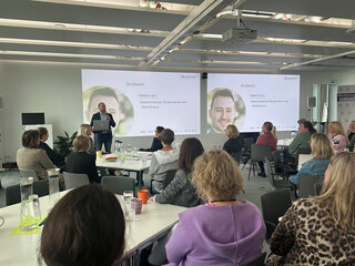 Das Bild zeigt eine Präsentation in einem modernen Raum. Ein Mann spricht vor einer Gruppe von Zuhörern, die gespannt zuhören.