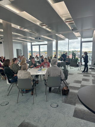 Das Bild zeigt eine Gruppe von Menschen in einem modernen Raum. Sie sitzen an Tischen und scheinen an einer Diskussion oder Präsentation teilzunehmen.