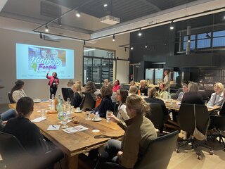 Das Bild zeigt eine Gruppe von Frauen in einem modernen Raum, die an einem Mentoring-Event teilnehmen. Eine Frau präsentiert auf einer Leinwand.