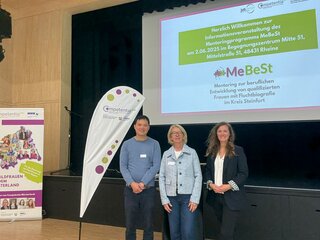 Das Bild zeigt eine Willkommensveranstaltung des Mentorenprogramms MeBeSt in Rheine. Drei Personen stehen vor einer Bühne mit einem Info-Stand.