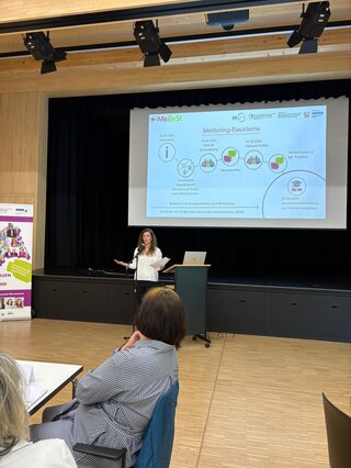 Eine Frau steht auf einer Bühne und präsentiert Informationen zu einem Mentoring-Programm. Im Hintergrund sind Diagramme und Daten zu sehen.