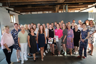 Auf dem Bild sind viele Frauen versammelt, die lächeln und feiern. Sie stehen in einem hellen Raum mit Holzdecke und präsentieren Urkunden.