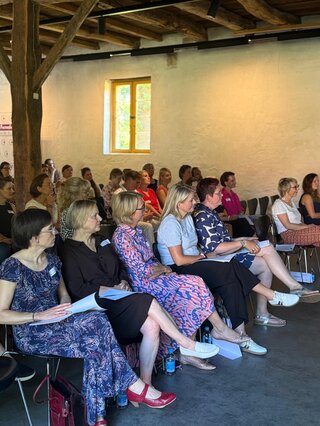 Eine Gruppe von Menschen sitzt aufmerksam in einem Raum mit hölzernen Balken. Einige haben Notizen, alle hören einer Präsentation zu.