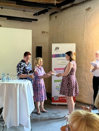 Das Bild zeigt eine Veranstaltung mit mehreren Personen, die einander Gratulationen aussprechen. Im Hintergrund sind Informationsbanner und Tischware.