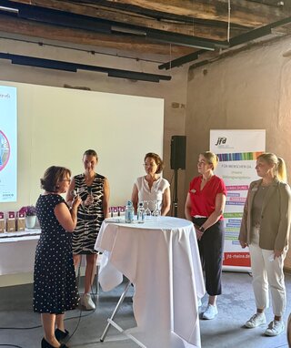 Auf dem Bild sind fünf Frauen zu sehen, die an einem Tisch stehen und diskutieren. Hintergrund ist ein heller Raum mit Werbematerialien.