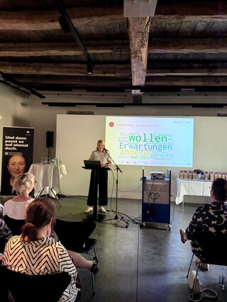 Die Präsentation zeigt eine Frau am Rednerpult, während ein Bildschirm mit Textinhalten im Hintergrund sichtbar ist. Zuhörer sitzen im Raum.