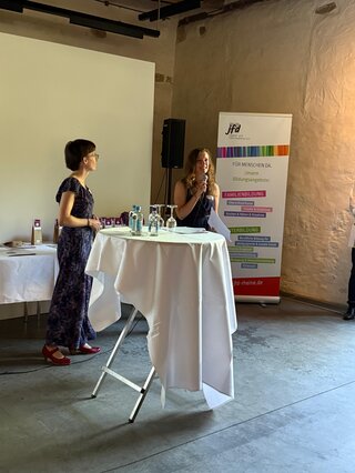 Zwei Frauen stehen an einem Tisch in einem Innenraum. Eine spricht, während die andere zuhört. Im Hintergrund ist ein Banner mit Informationen zu sehen.