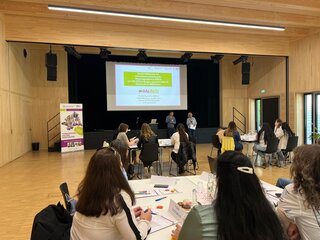 Das Bild zeigt eine Präsentation in einem hellen Raum mit Sitzplätzen. Teilnehmer hören aufmerksam zu, während zwei Personen auf der Bühne stehen.