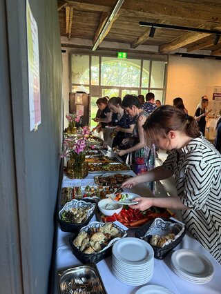 Eine Gruppe von Menschen steht an einem Buffet, während sie sich verschiedene Speisen auf Teller nehmen. Der Raum hat eine einladende Atmosphäre.