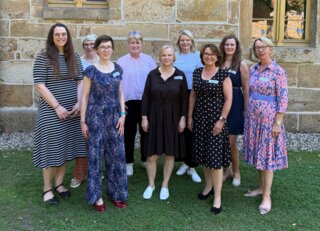 Eine Gruppe von Frauen steht lächelnd vor einer alten Steinmauer. Sie tragen verschiedene Kleider und wirken freundlich und entspannt.