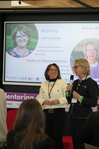 Das Bild zeigt zwei Frauen, die auf einer Veranstaltung sprechen. Eine begrüßt die andere, während die Teilnehmerinnen aufmerksam zuhören.