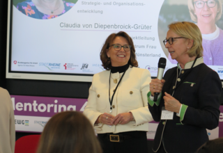 Das Bild zeigt zwei Frauen, die auf einer Veranstaltung sprechen. Eine begrüßt die andere, während die Teilnehmerinnen aufmerksam zuhören.