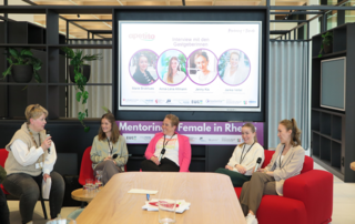 Das Bild zeigt eine Panel-Diskussion mit vier Frauen. Sie sprechen auf einer Bühne, während eine Moderatorin Fragen stellt. Im Hintergrund sind Präsentationen sichtbar.