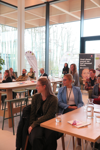Das Bild zeigt eine Gruppe von Frauen, die in einem modernen Raum an Tischen sitzen. Sie hören aufmerksam zu und scheinen an einer Präsentation teilzunehmen.