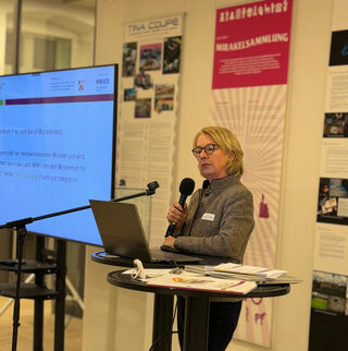 Eine Frau steht an einem Stehschreibtisch und spricht mit einem Laptop vor einem Bildschirm. Im Hintergrund sind Plakate und Informationen sichtbar.