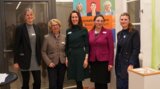 Das Bild zeigt fünf Frauen, die zusammen in einer Ausstellung oder Veranstaltung stehen. Im Hintergrund ist ein Banner mit Informationen sichtbar.