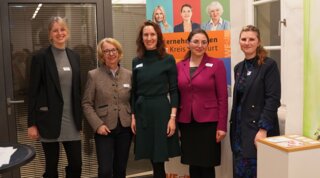 Das Bild zeigt fünf Frauen, die zusammen in einer Ausstellung oder Veranstaltung stehen. Im Hintergrund ist ein Banner mit Informationen sichtbar.
