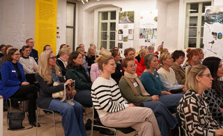 Das Bild zeigt eine Gruppe von Menschen, die aufmerksam in einem Raum sitzen. Der Raum ist hell und gut beleuchtet, mit informativen Ausstellungen im Hintergrund.