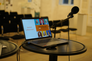 Das Bild zeigt einen Laptop auf einem Tisch mit einer Willkommensnachricht und einer Mikrofonaufnahme. Im Hintergrund sind leere Stühle zu sehen.