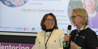 Das Bild zeigt zwei Frauen, die auf einer Veranstaltung sprechen. Eine Frau begrüßt die Anwesenden, während die andere in einem Mikrofon spricht.