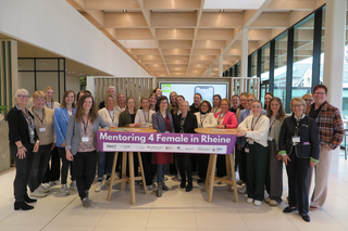Eine Gruppe von Frauen steht zusammen in einem modernen Raum und hält ein Banner mit der Aufschrift „Mentoring 4 Female in Rheine“.