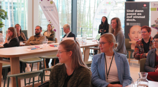 Eine Gruppe von Menschen sitzt in einem hellen Raum und verfolgt aufmerksam eine Präsentation. Tische mit Wasserkrügen sind vorhanden.