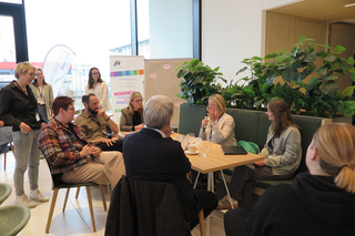 Eine Gruppe von Menschen sitzt in einem modernen Raum, diskutiert an einem Tisch und genießt Getränke. Pflanzen verzieren die Umgebung.