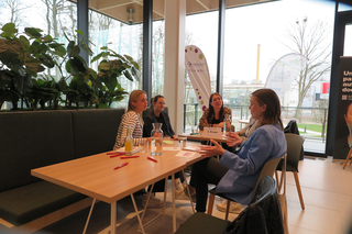 Vier Frauen sitzen an einem Tisch in einem modernen Raum. Sie unterhalten sich lebhaft, umgeben von Pflanzen und großen Fenstern mit Blick nach draußen.