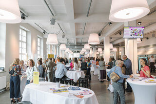 Das Bild zeigt eine lebhafte Veranstaltung mit vielen Menschen, die an Stehtischen plaudern. Helle Räume und große Fenster sorgen für eine freundliche Atmosphäre.