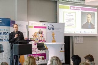 Eine Person spricht am Rednerpult auf einer Veranstaltung. Im Hintergrund sind Banner und eine Präsentation mit einem Porträt zu sehen.