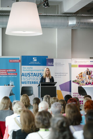 Eine Frau hält eine Rede vor Publikum. Im Hintergrund sind Banner mit Informationen zu Marketing und Weiterbildung sichtbar.