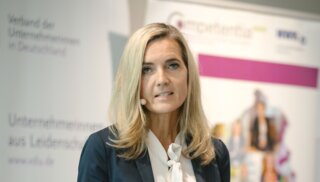 Eine Geschäftsführerin spricht am Rednerpult. Im Hintergrund sind informative Plakate und ein Logo zu sehen. Sie wirkt professionell und engagiert.